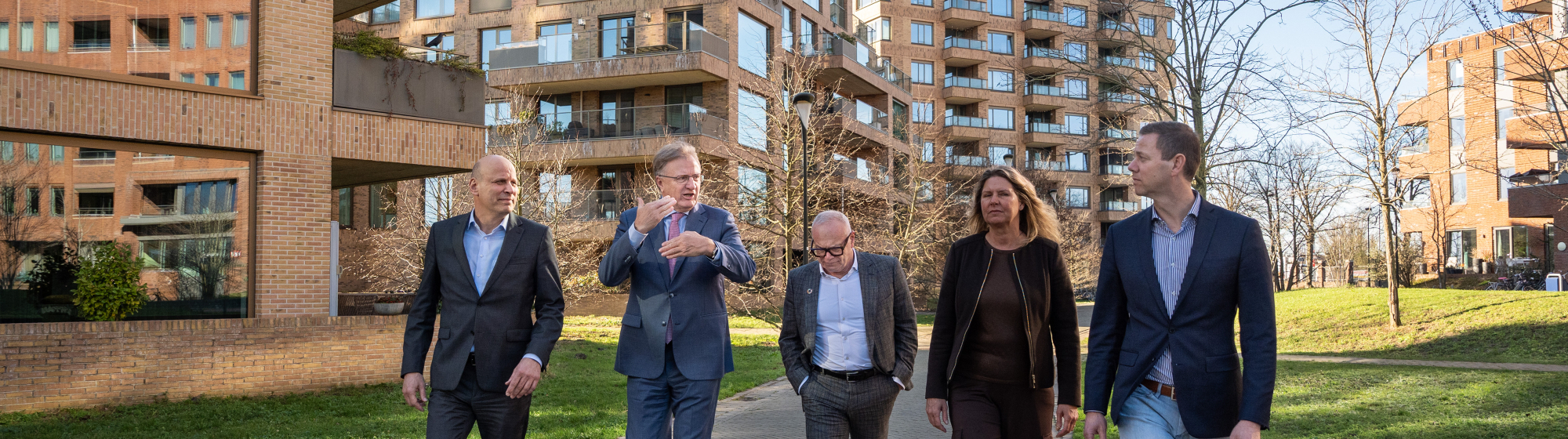 De bestuurders van de samenwerkingspartijen lopen naast elkaar tussen flatgebouwen, omgeven door groen en grasvelden.