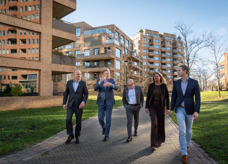 De bestuurders van de samenwerkingspartijen lopen naast elkaar tussen flatgebouwen, omgeven door groen en grasvelden.