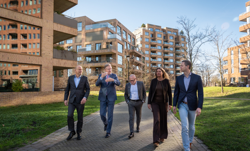 De bestuurders van de samenwerkingspartijen lopen naast elkaar tussen flatgebouwen, omgeven door groen en grasvelden.