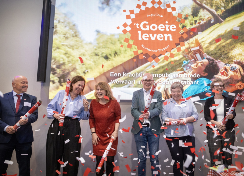 Feestelijk moment tijdens Regio Deal startbijeenkomst bestuurders met confetti op het podium. Op de achtergrond een foto met het Regio Deal logo.