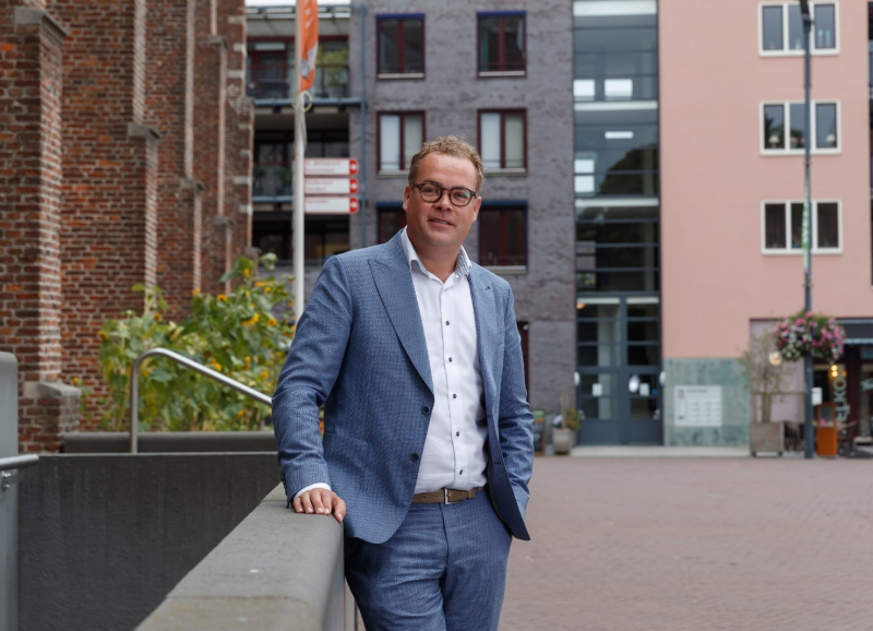 Erik Kiers buiten in stedelijke omgeving met moderne gebouwen en groene beplanting op de achtergrond.