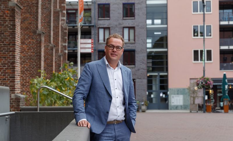 Erik Kiers buiten in stedelijke omgeving met moderne gebouwen en groene beplanting op de achtergrond.