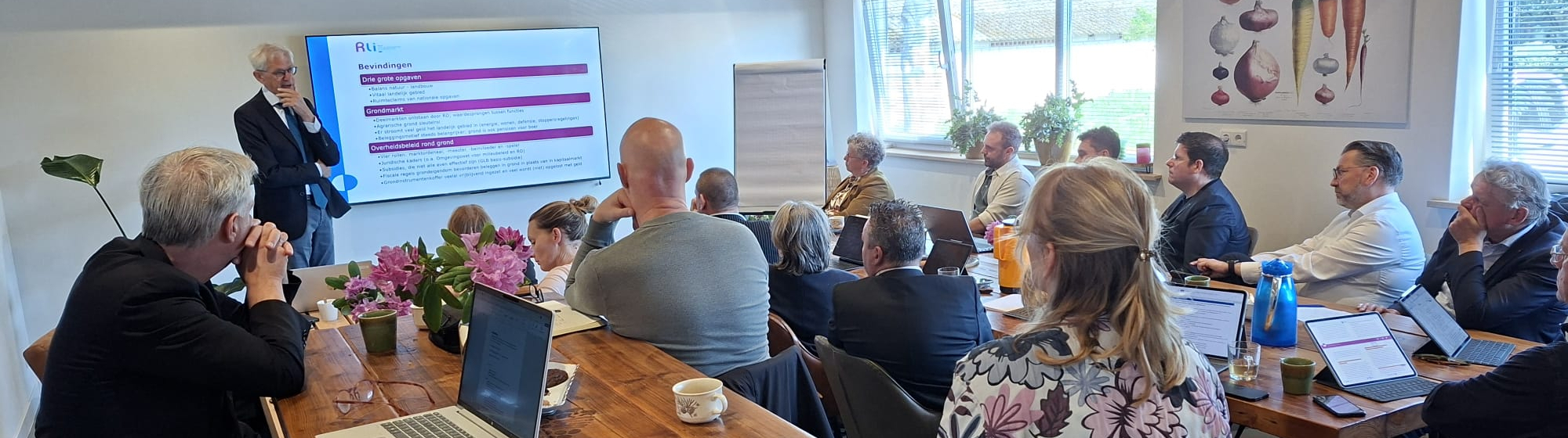Wethouders uit Noordoost-Brabant luisteren in een zaaltje naar een presentatie van Krijn Poppe vóór een scherm.