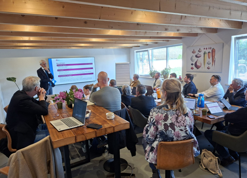 Wethouders uit Noordoost-Brabant luisteren in een zaaltje naar een presentatie van Krijn Poppe vóór een scherm.