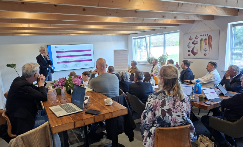 Wethouders uit Noordoost-Brabant luisteren in een zaaltje naar een presentatie van Krijn Poppe vóór een scherm.