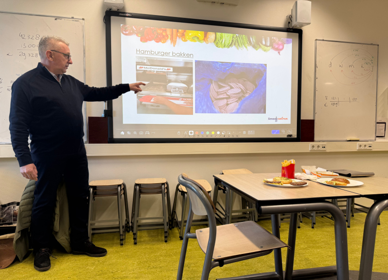 Martien Bos geeft een presentatie over hamburgerproductie en ingrediënten in een klaslokaal.