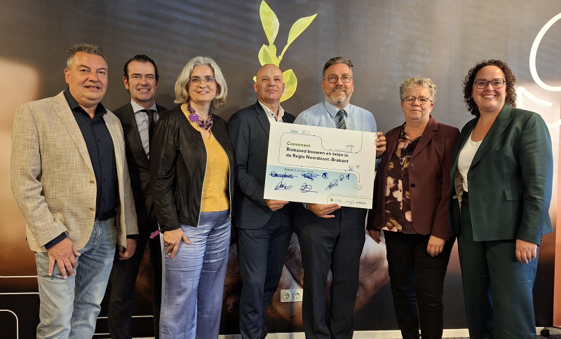 Zeven wethouders op naast elkaar op een rij vóór een afbeelding met een gewas, één houdt een bord met een convenant en handtekeningen in de hand.