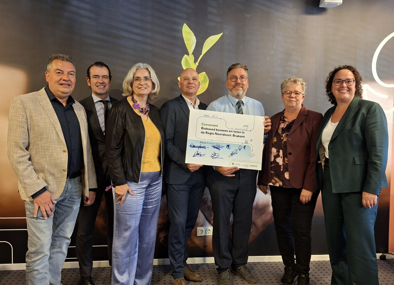 Zeven wethouders op naast elkaar op een rij vóór een afbeelding met een gewas, één houdt een bord met een convenant en handtekeningen in de hand.