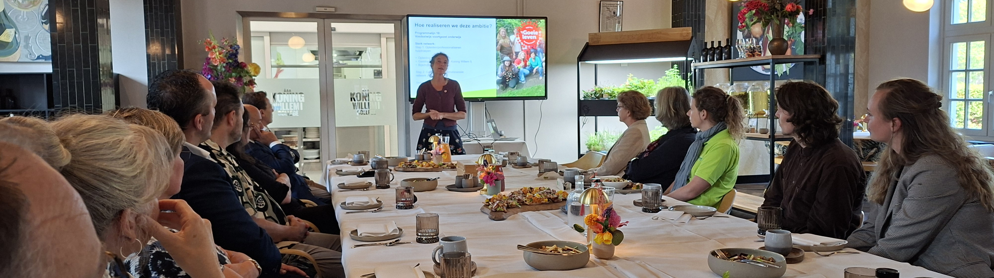 Groep mensen rond een lange, gedekte tafel tijdens een bijeenkomst, met voor in de ruimte een spreker en een presentatiescherm.