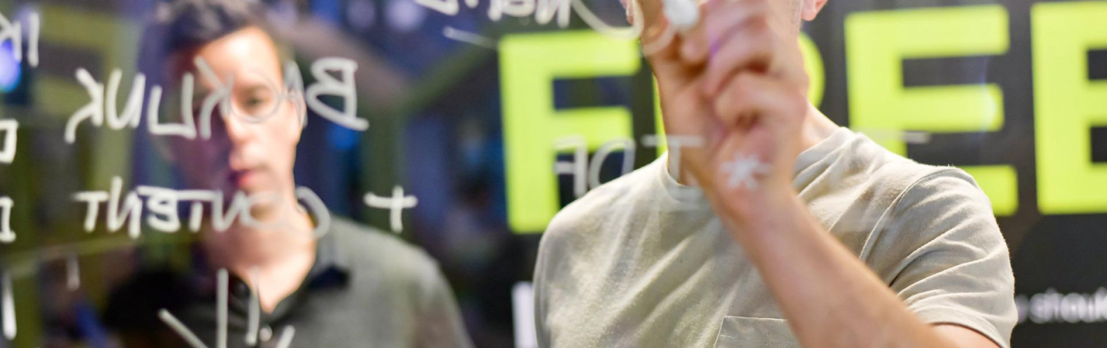 Foto van jonge ondernemer die met een stift op een glasplaat schrijft, met een oudere man op de achtergrond