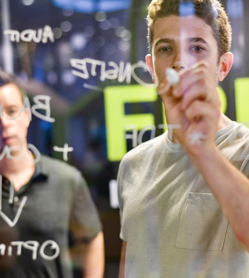 Foto van jonge ondernemer die met een stift op een glasplaat schrijft, met een oudere man op de achtergrond