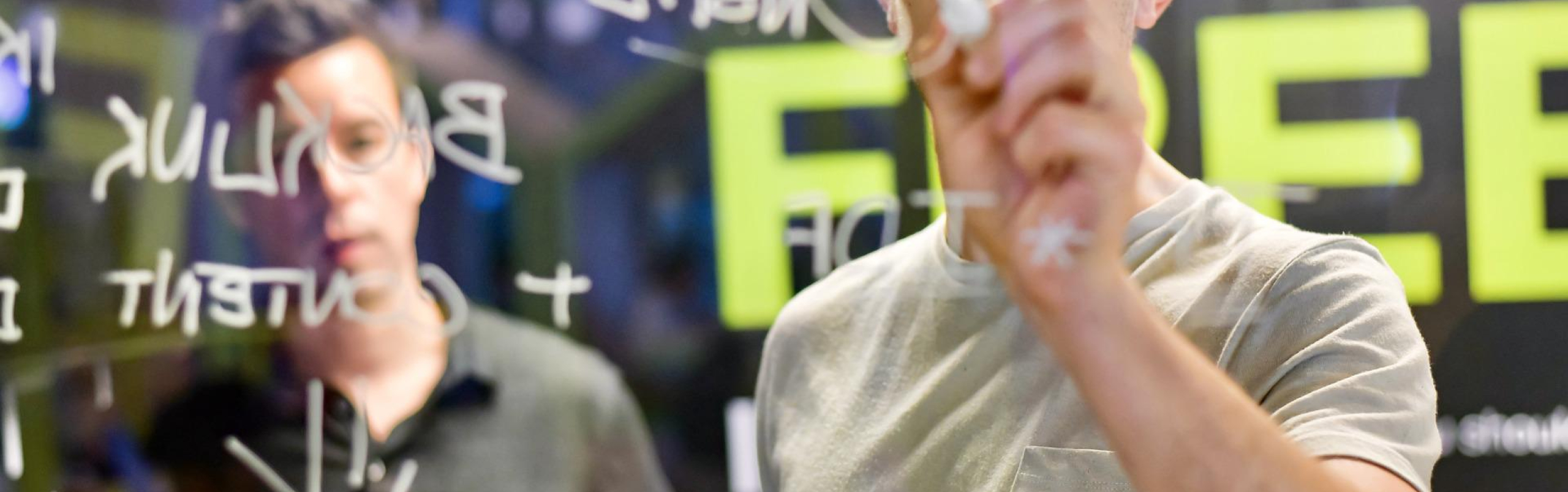 Foto van jonge ondernemer die met een stift op een glasplaat schrijft, met een oudere man op de achtergrond