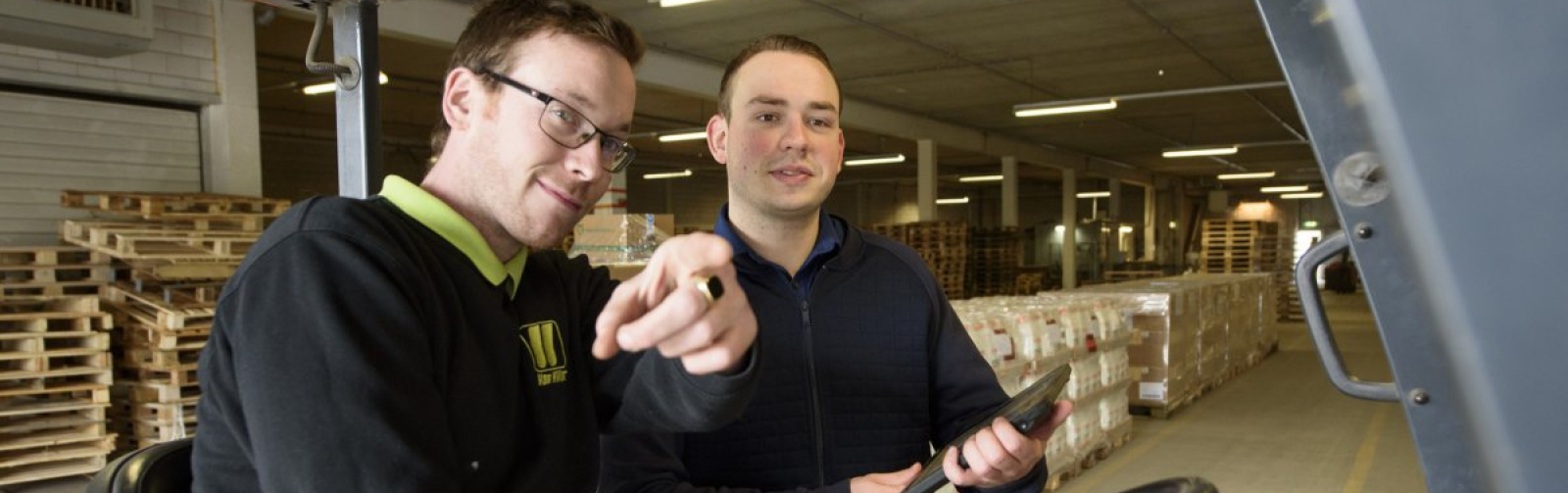 Foto van twee werknemers op en naast een vorkheftruck in een magazijn opls