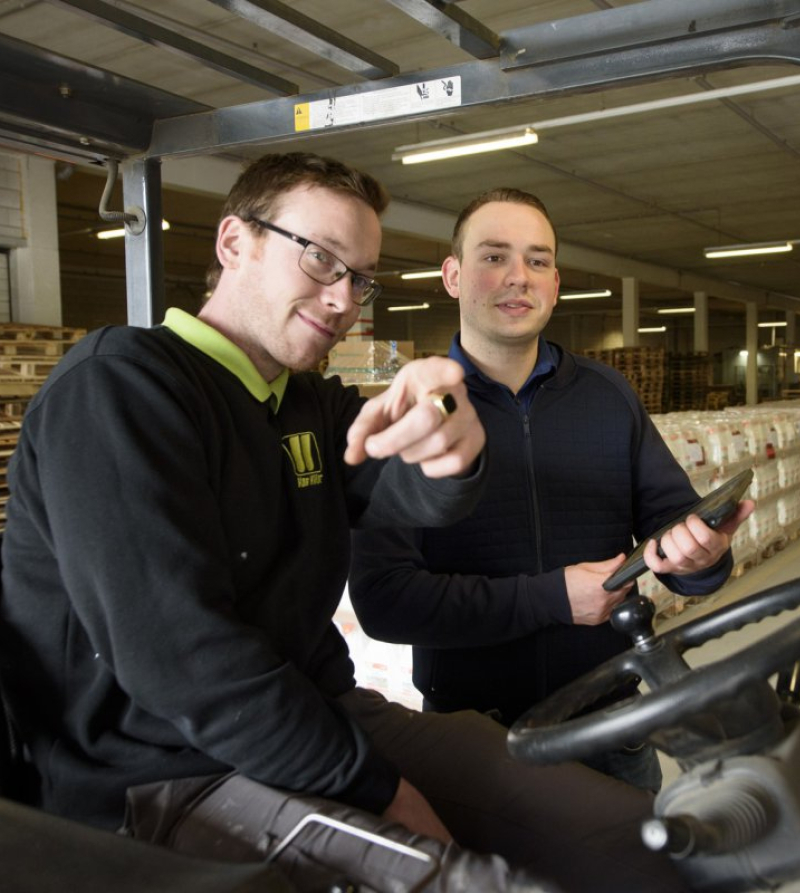 Foto van twee werknemers op en naast een vorkheftruck in een magazijn opls