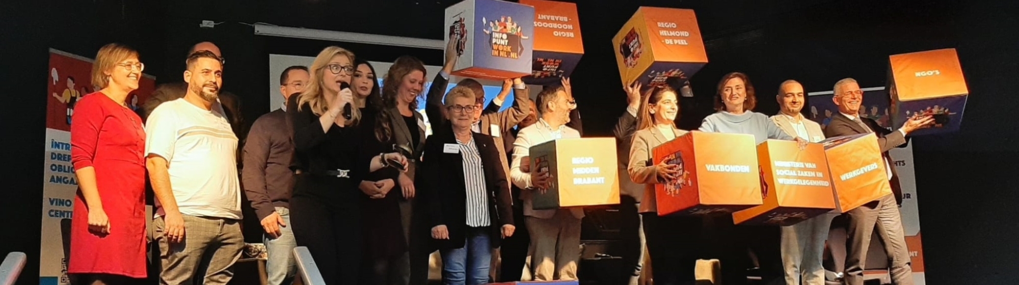 Groep mensen op een podium poseert samen met grote gekleurde blokken met teksten en illustraties.