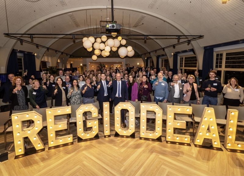 Groepsfoto van deelnemers aan het Regio Deal slotevent achter het woord Regio Deal in grote lichtgevende letters