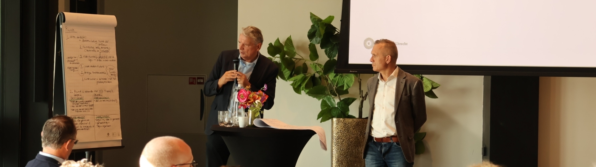 Twee sprekers, waaronder Marcel Boogers, presenteren voor een groep aanwezigen in een zaal, met een groot projectiescherm en flip-over op de achtergrond.