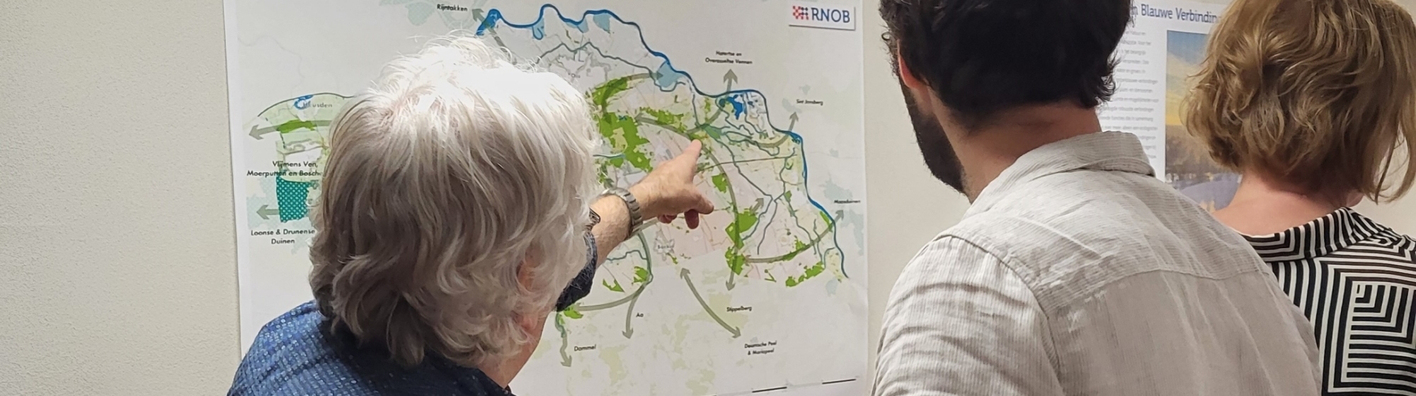 Drie deelnemers van de netwerkbijeenkomst staan een muur met een landkaart 