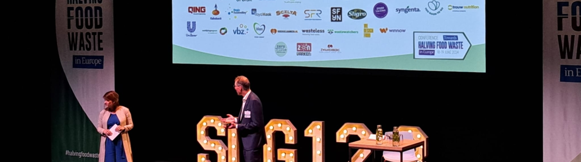 Twee sprekers op het podium van de bijeenkomst onder een slide met de logo's van de partners