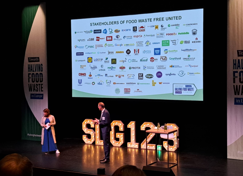 Twee sprekers op het podium van de bijeenkomst onder een slide met de logo's van de partners