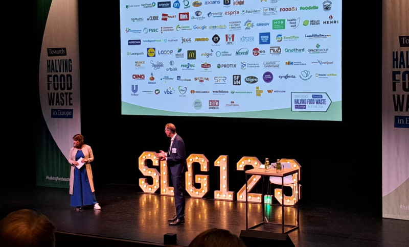 Twee sprekers op het podium van de bijeenkomst onder een slide met de logo's van de partners