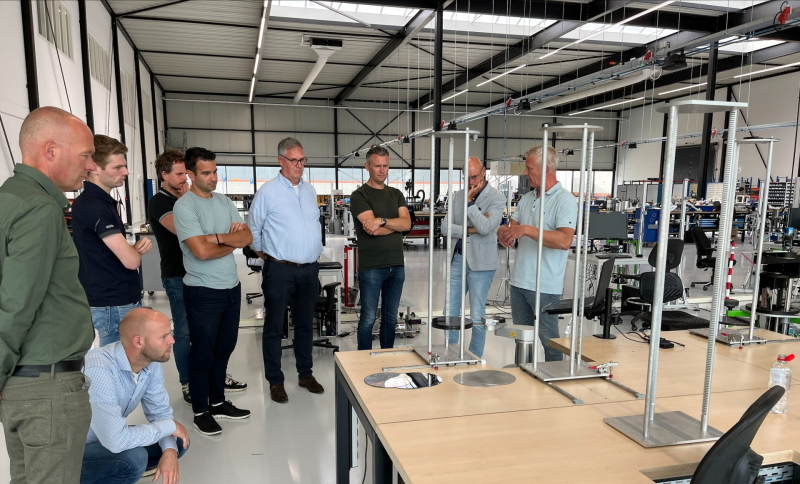 Groep mannen in een werkplaats luistert naar een presentatie rond metalen frames bij werktafels
