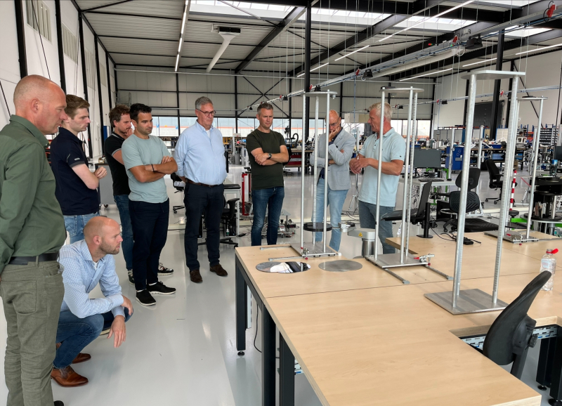 Groep mannen in een werkplaats luistert naar een presentatie rond metalen frames bij werktafels