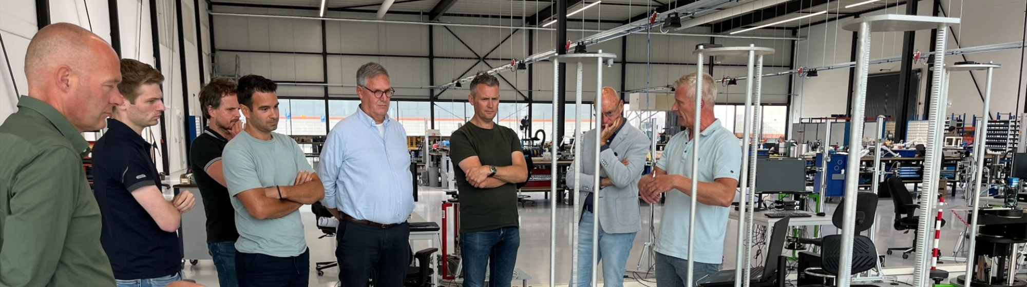 Groep mannen in een werkplaats luistert naar een presentatie rond metalen frames bij werktafels