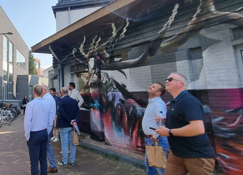 Groep bestuurders bekijkt een grote muurschildering van een hert op de gevel van een gebouw.
