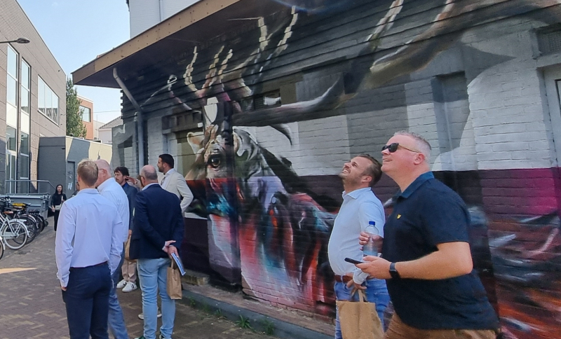 Groep bestuurders bekijkt een grote muurschildering van een hert op de gevel van een gebouw.