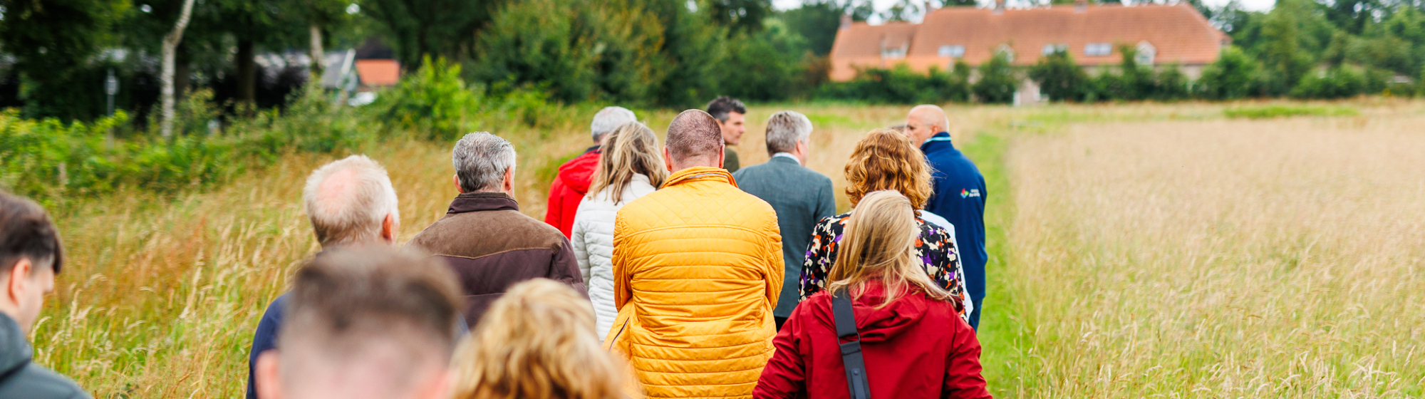 Groep mensen wandelt over een smal pad door een veld richting een boerderij, op een bewolkte dag.