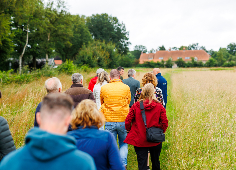 Groep mensen wandelt over een smal pad door een veld richting een boerderij, op een bewolkte dag.