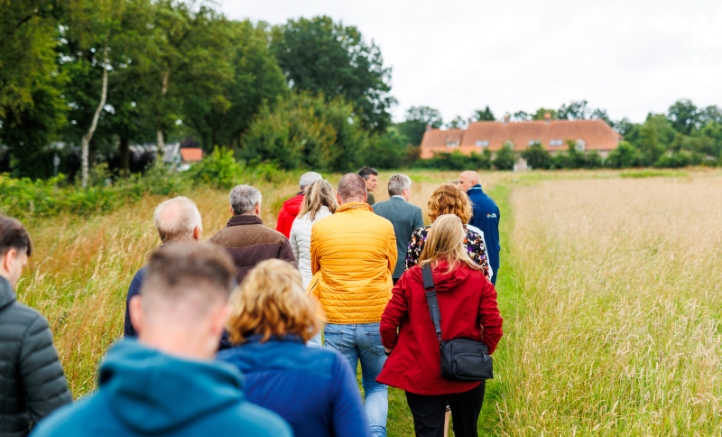 Groep mensen wandelt over een smal pad door een veld richting een boerderij, op een bewolkte dag.