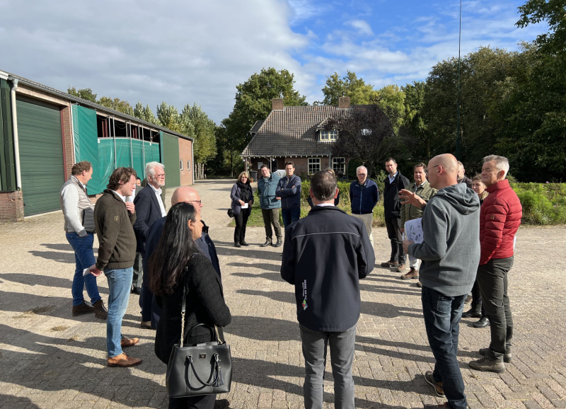 Groep mensen staat in een kring bij elkaar op een versteerd stuk grond buiten bij een boerderij op een zonnige dag
