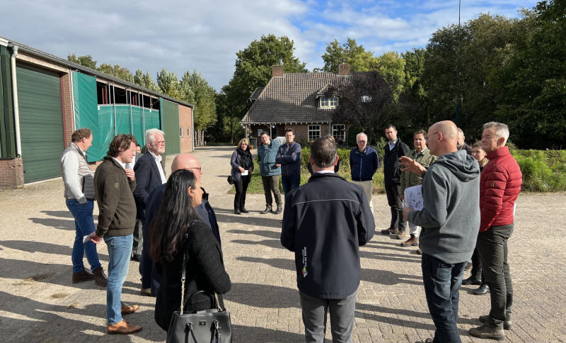 Groep mensen staat in een kring bij elkaar op een versteerd stuk grond buiten bij een boerderij op een zonnige dag