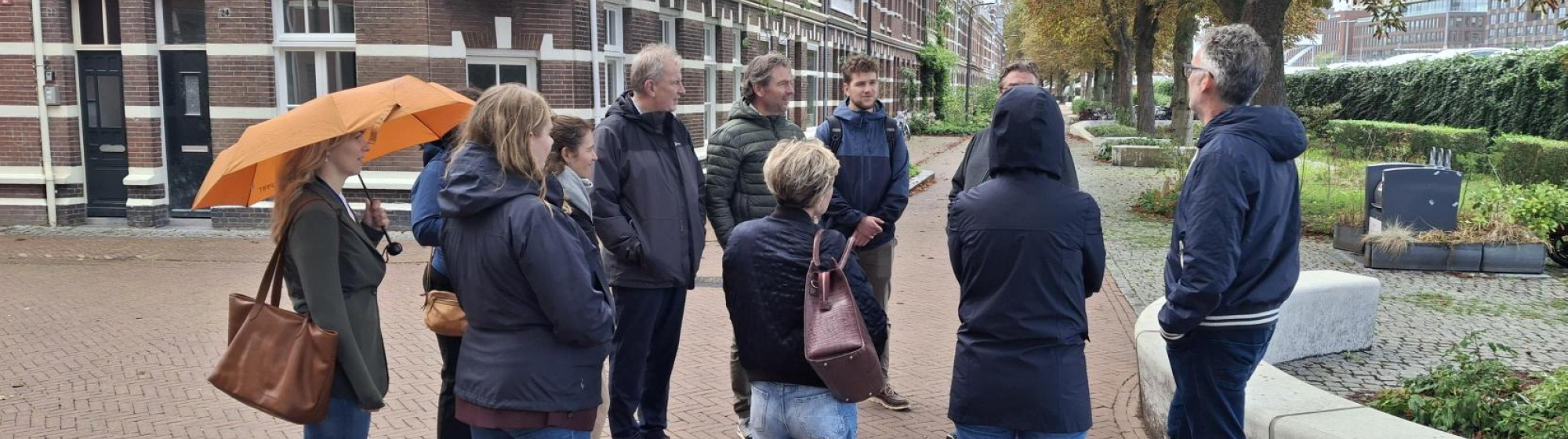 Een groep mensen in miezerregen met elkaar in gesprek op een plein in Den Bosch met huizen en bomen