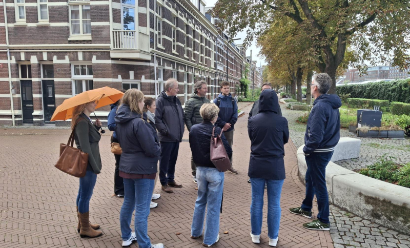 Een groep mensen in miezerregen met elkaar in gesprek op een plein in Den Bosch met huizen en bomen
