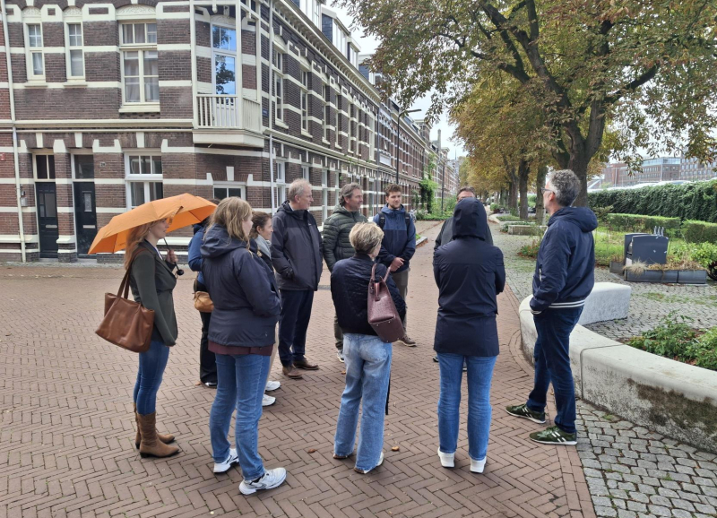 Een groep mensen in miezerregen met elkaar in gesprek op een plein in Den Bosch met huizen en bomen