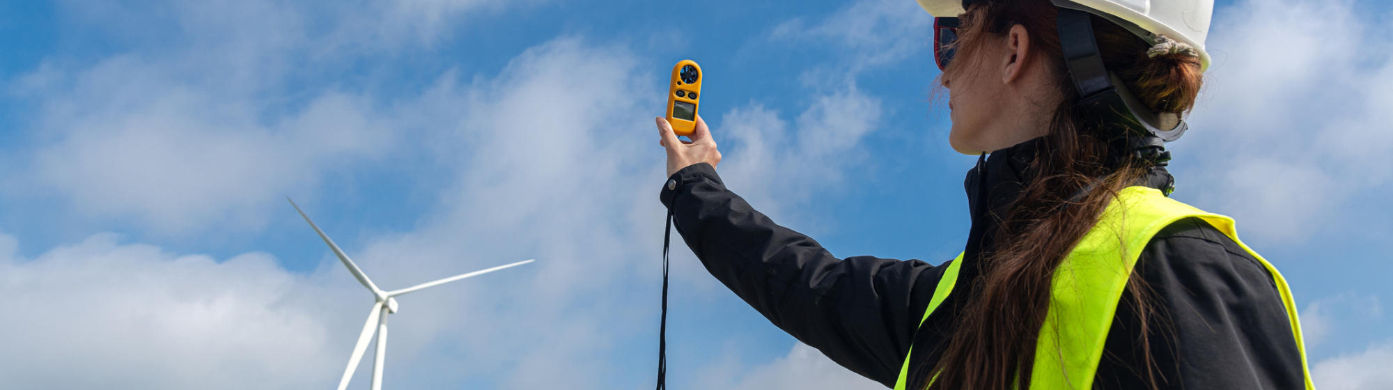 Vrouwelijke ingenieur met veiligheidshelm en reflecterend vest voert metingen uit met meetapparatuur bij een windmolenpark.
