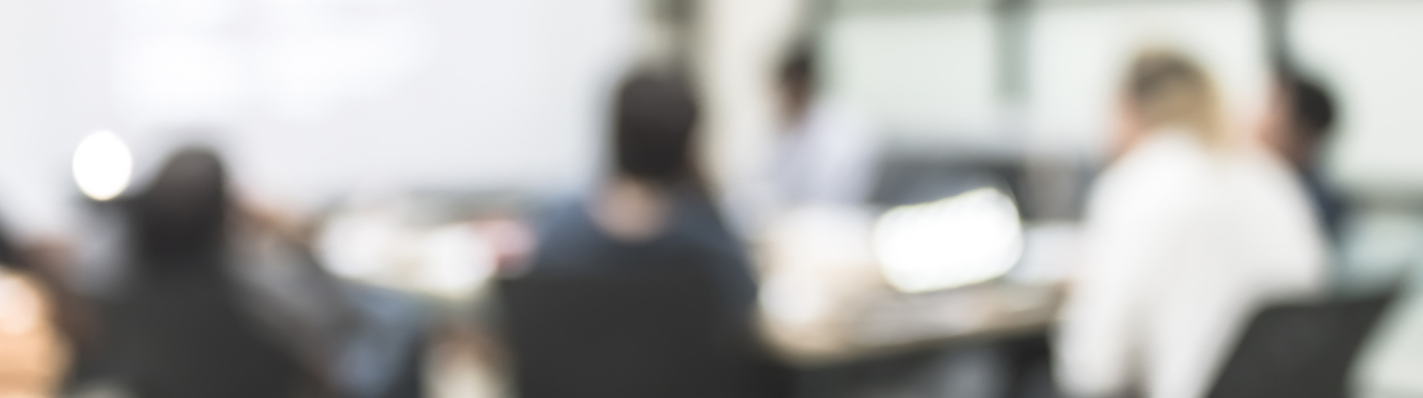 Vervaagde scène van een vergadering in een kantoorruimte, met mensen rond een conferentietafel, laptops en documenten voor zich, en een presentatie op een scherm.