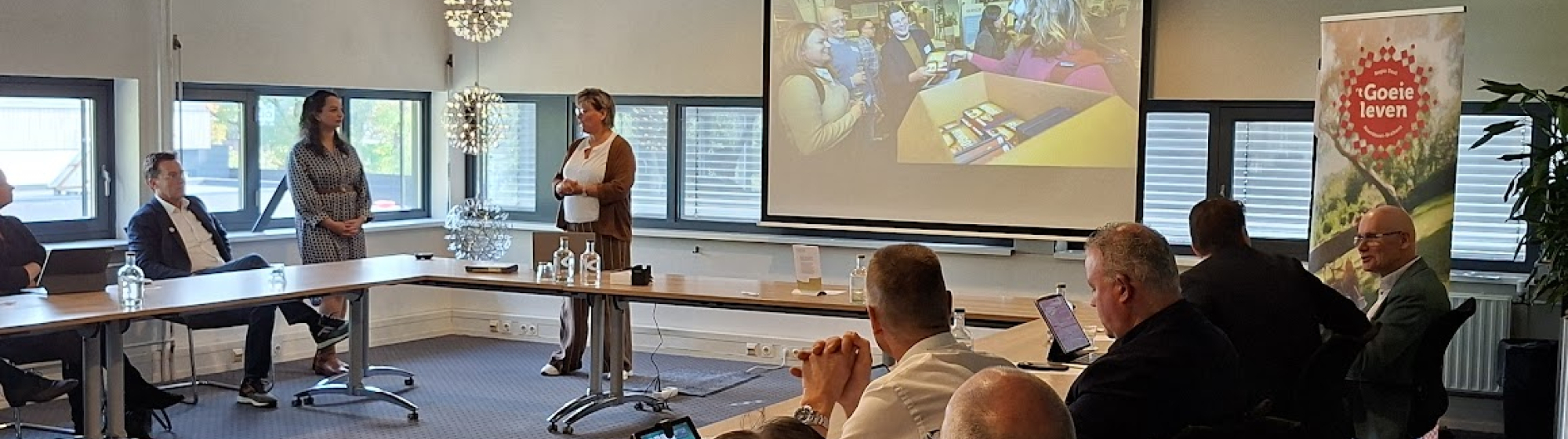 Presentatie in vergaderzaal met wethouders aan een lange tafel, een spreker voor een scherm met kleurrijke dia, en banner met Regio Deal 't Goeie leven-logo op de achtergrond.