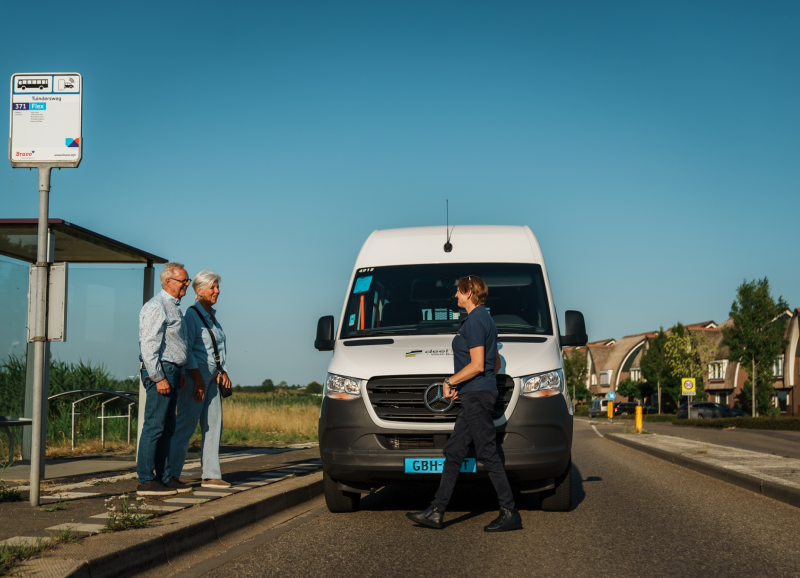 Witte BravoFlex bus bij bushalte in landelijke omgeving, met twee reizigers en chauffeur voor de bus.