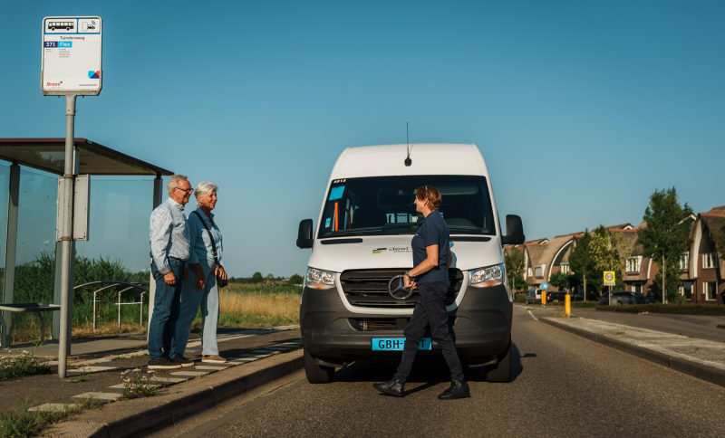 Witte BravoFlex bus bij bushalte in landelijke omgeving, met twee reizigers en chauffeur voor de bus.