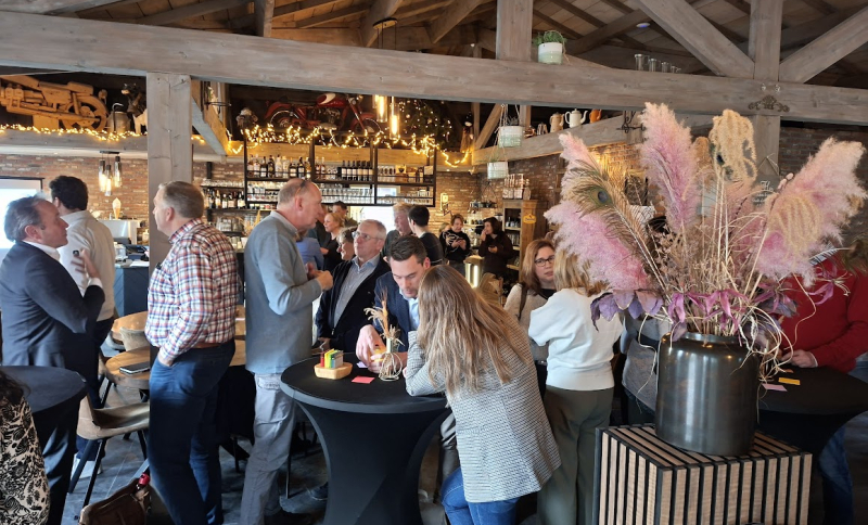 Deelnemers van de bijeenkomst in een tot eventlocatie omgebouwde boerderij met houten interieur, statafels en decoratieve pampaspluimen op de voorgrond.