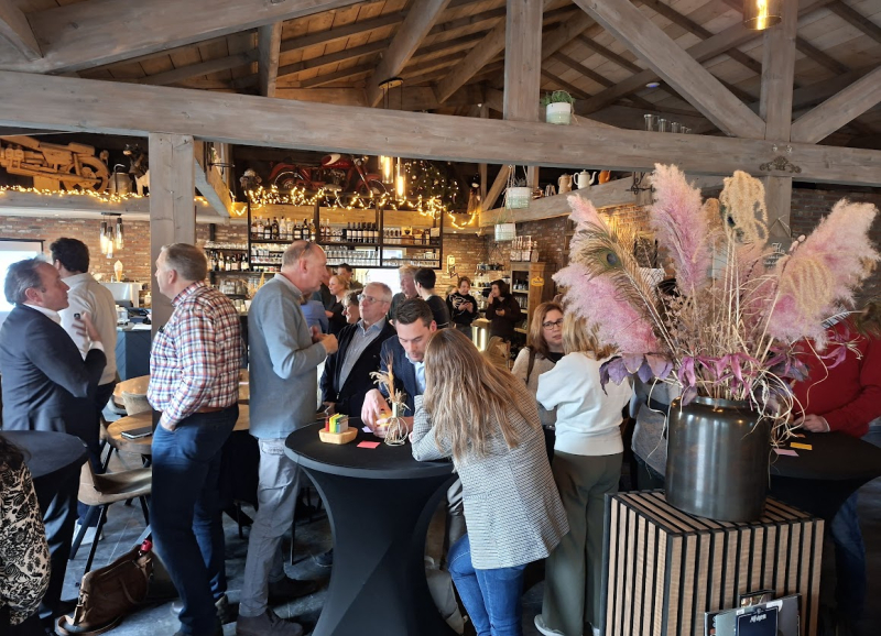 Deelnemers van de bijeenkomst in een tot eventlocatie omgebouwde boerderij met houten interieur, statafels en decoratieve pampaspluimen op de voorgrond.