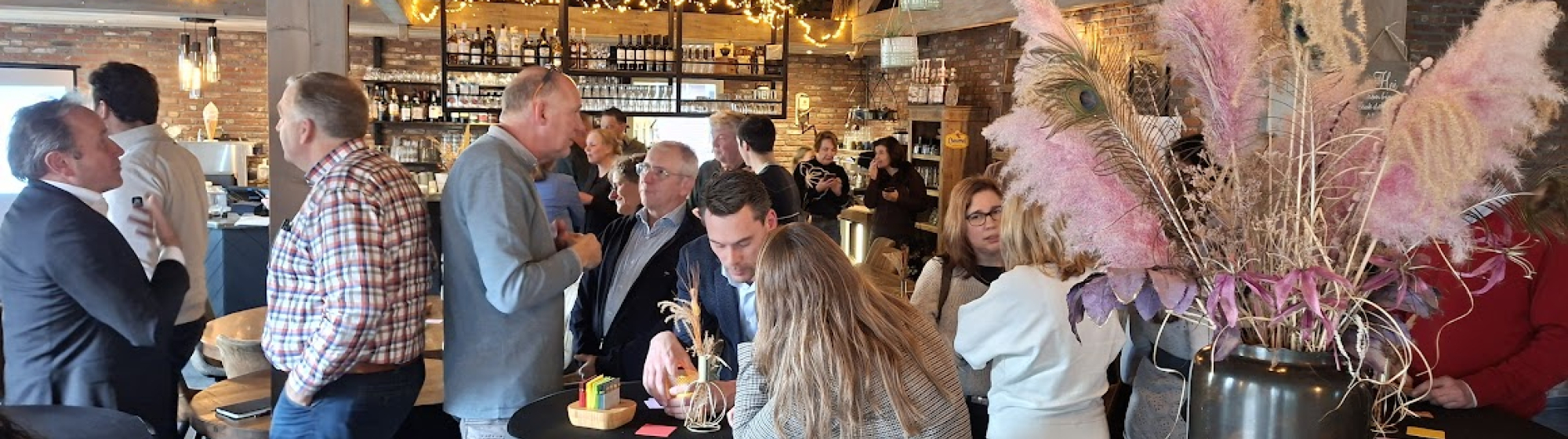 Deelnemers van de bijeenkomst in een tot eventlocatie omgebouwde boerderij met houten interieur, statafels en decoratieve pampaspluimen op de voorgrond.