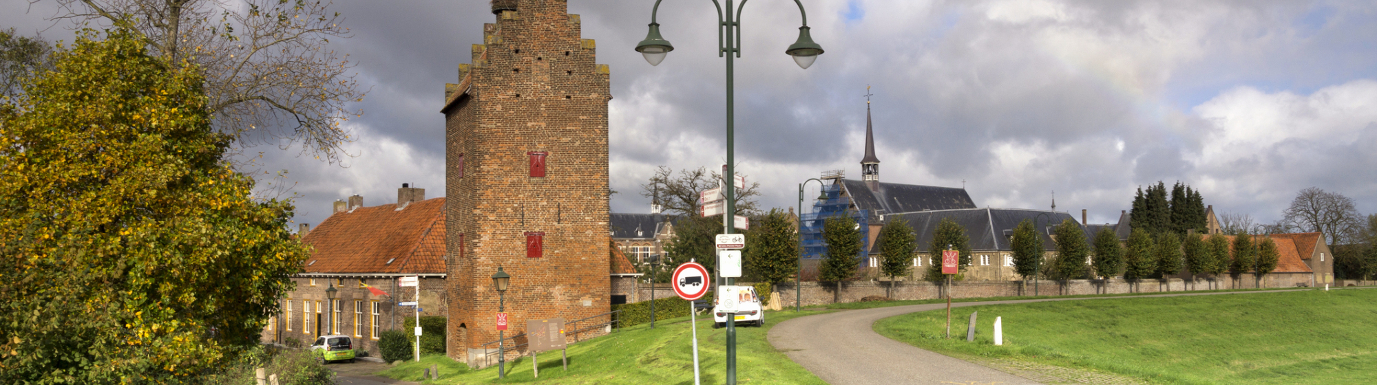 Historische toren en kerk in groen met een weg ervoor in een Noordoost-Brabants dorp met bewolkte lucht