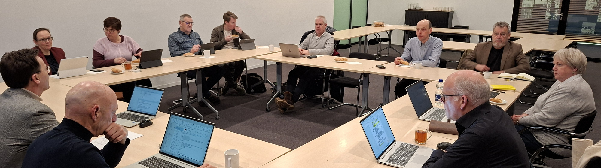 Overlegsessie met raadsleden aan vergadertafels die samenwerken en overleggen met laptops en notities.