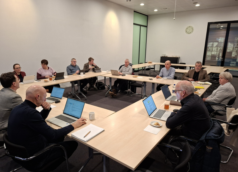 Overlegsessie met raadsleden aan vergadertafels die samenwerken en overleggen met laptops en notities.