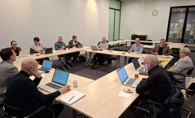 Overlegsessie met raadsleden aan vergadertafels die samenwerken en overleggen met laptops en notities.